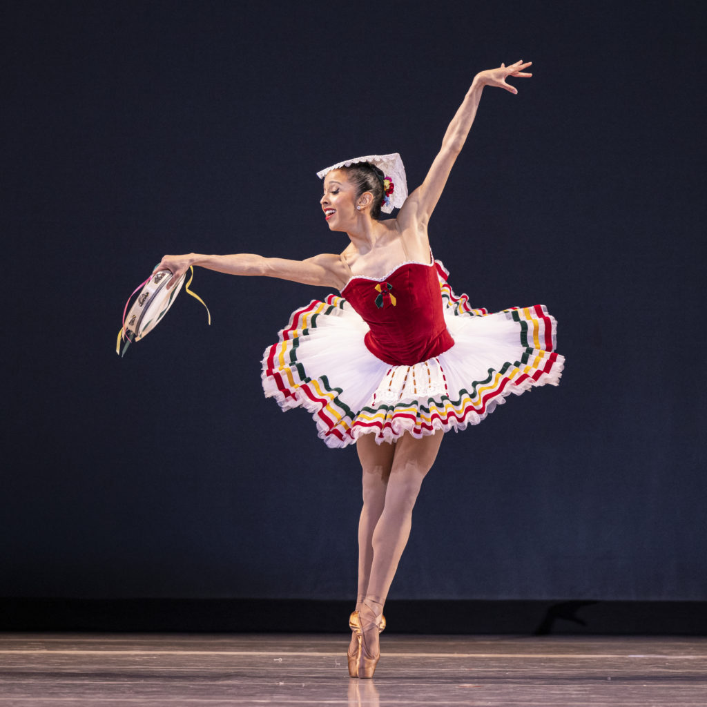 Jenna Rae Herrera stands in sous sous in a big white and red tutu. She holds a tambourine in her left arm and her right arm is raised overhead in open fourth.  
