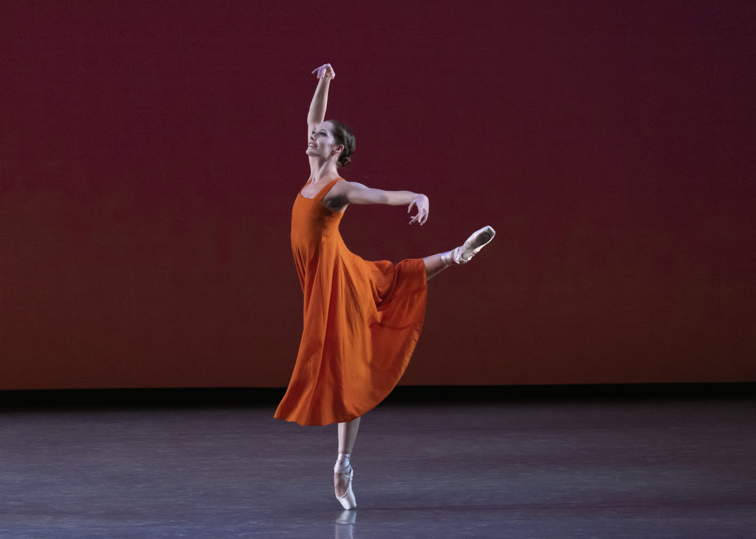 Unity Phelan is shown in profile on pointe raising her right leg behind her in attitude. She holds her right arm high and her left arm out to the side and looks up towards her raised hand. She wears a bright orange dress and dances onstage in front of an orange-brown backdrop.