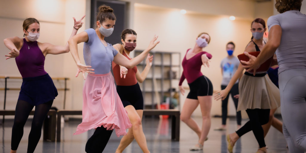 A group of female ballerinas in dance clothing twist their bodies in motion during a rehearsal in a ballet studio.