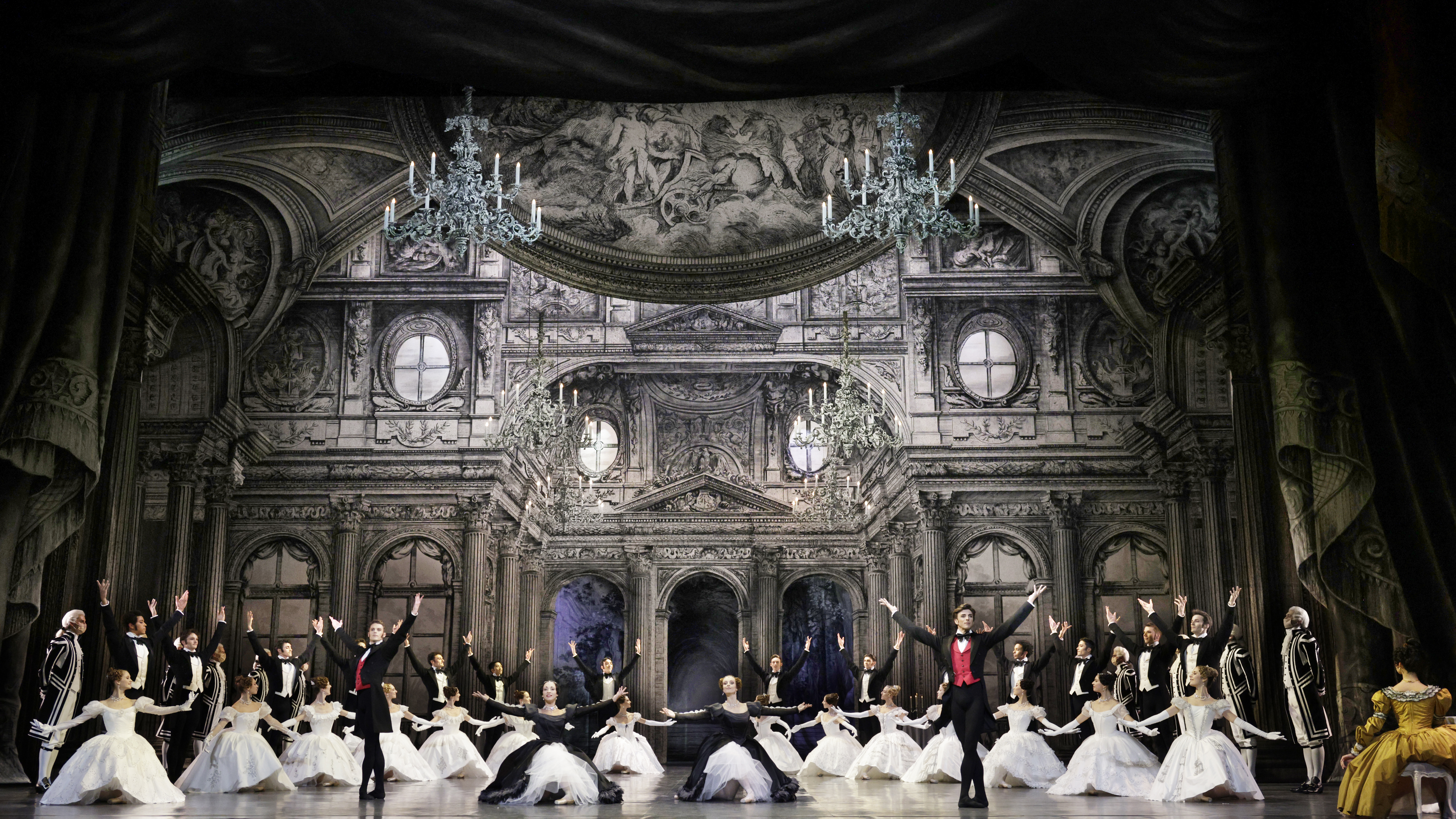 A large company of dancers pose in front of an elaborate ballroom set onstage. Four soloists form a line at the front of the stage, the women kneeling in between the two standing men. Behind them, 14 dancers form a V-shape formation, the women kneeling and their male partners standing behind them with their arms up. All of the men wear tuxedo jackets, black tights and balck slippers, while the women wear long tutu dresses with puffed sleeves.