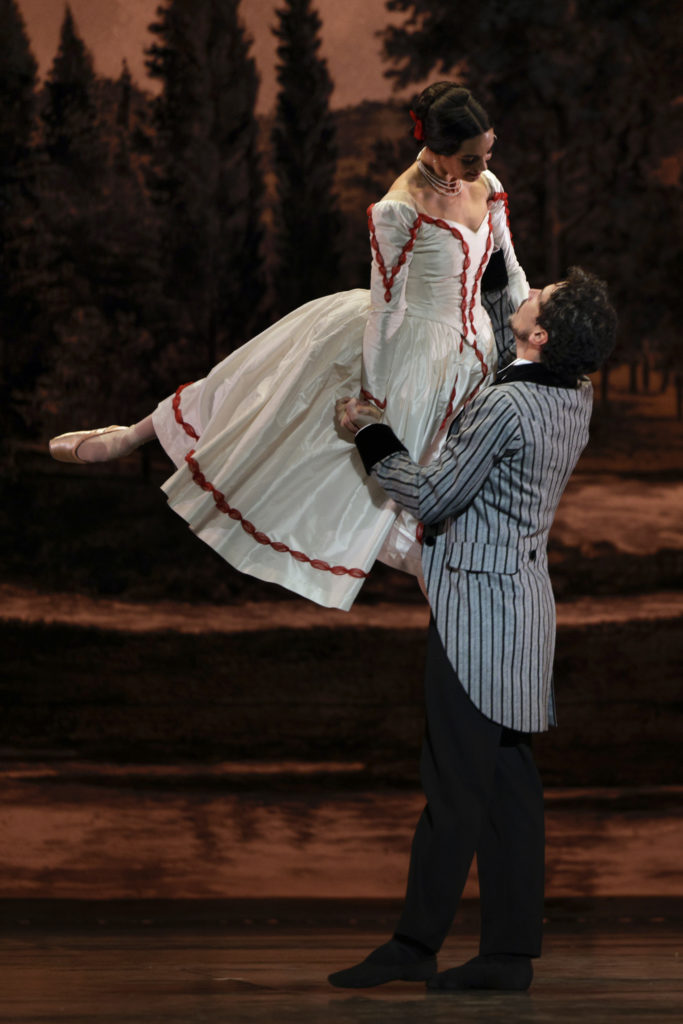 Stephan Bullion, dressed in a gray striped sport coat and black pants, lifts Amadine Albisson up by her left shoulder and right hand as she makes a stag position with her legs. She wears a long white gown with red trim and pointe shoes, and looks down at her partner with affection.