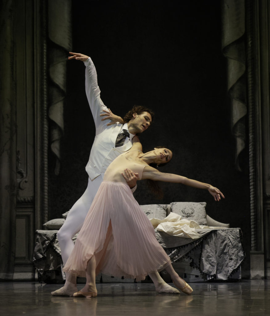 During a performance, Hugo Marchand, in a white vest, shirt and tights, holds Dorothée Gilbert around the waist as she swoons back in a lunge. She wears a light pink nightdress and pointe shoes.