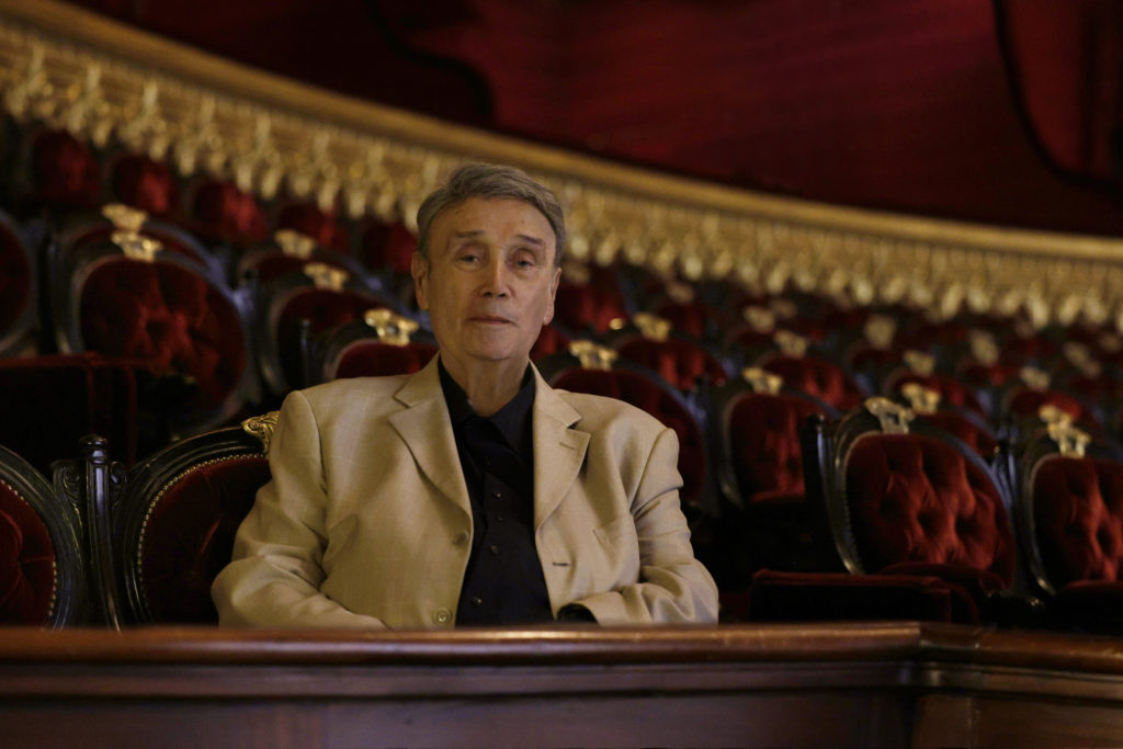 Pierre Lacotte, wearing a black turtleneck and tan sport coat, sits in a red velvet seat in the balcony of the Palais Garnier theater and smiles slightly to the camera.