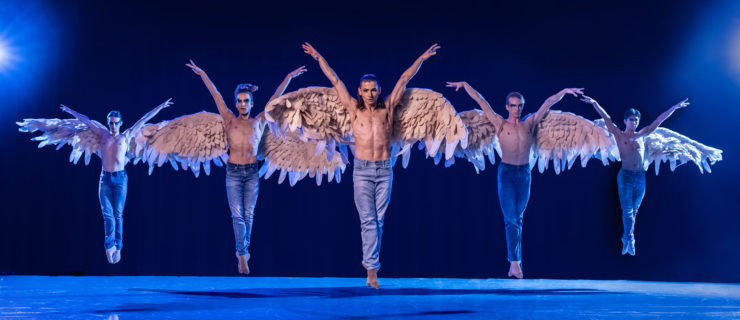 On a cooly lit stage, five male dancers wearing jeans and giant angel wings jump in sus-sous with their arms up in a V-shape. .