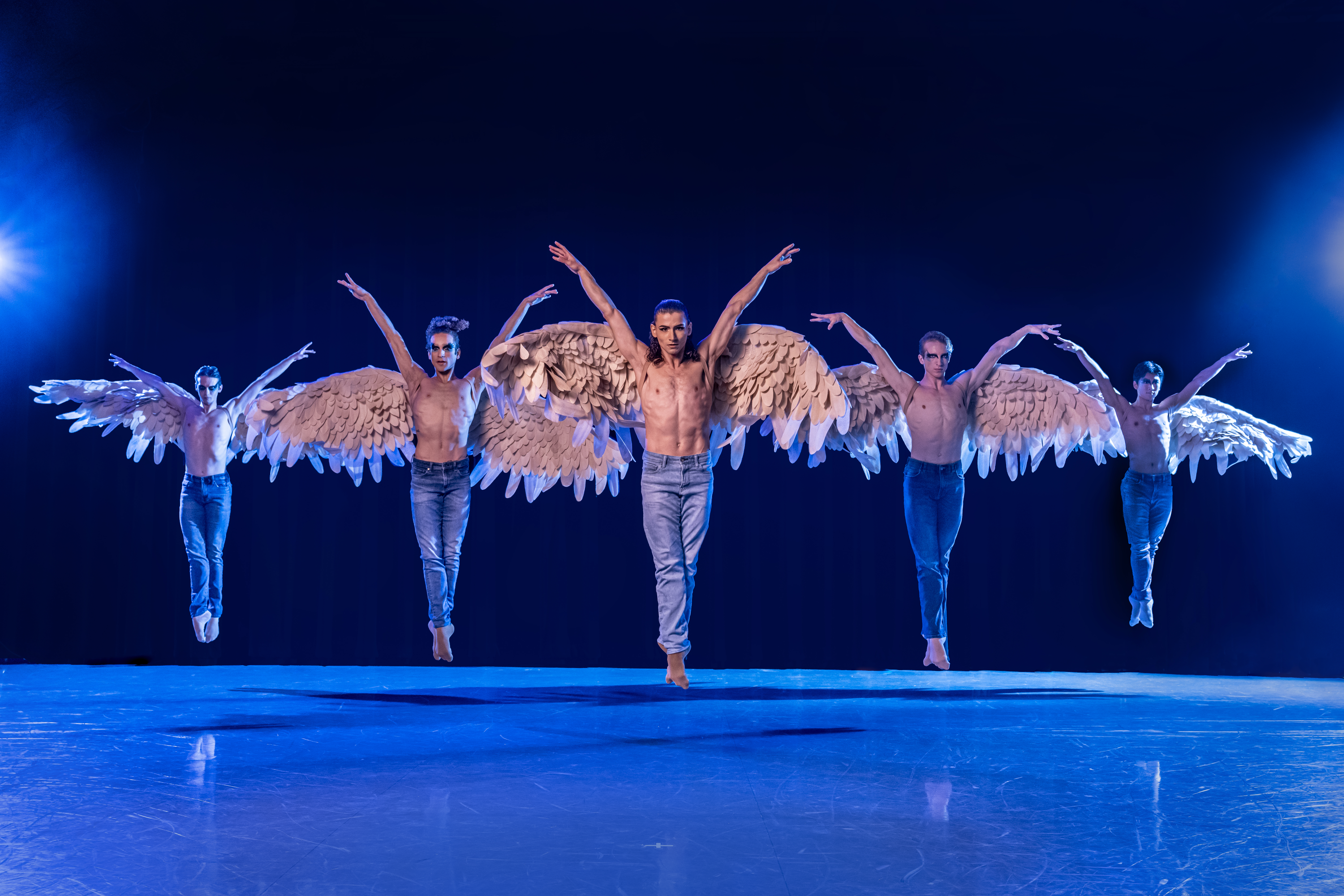 On a cooly lit stage, five male dancers wearing jeans and giant angel wings jump in sus-sous with their arms up in a V-shape. .