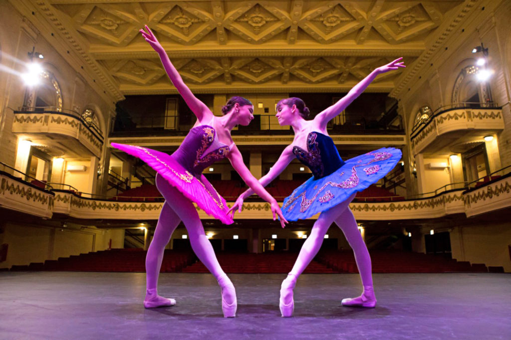 Two teenage female dancers in beautiful tutus face each other on a grand stage. They each stand in tendu croisé and bend over slightly so that their outstretched arms cross each other. The ballerina on the left wears a purple tutu, while the one on the right wears a blue tutu.