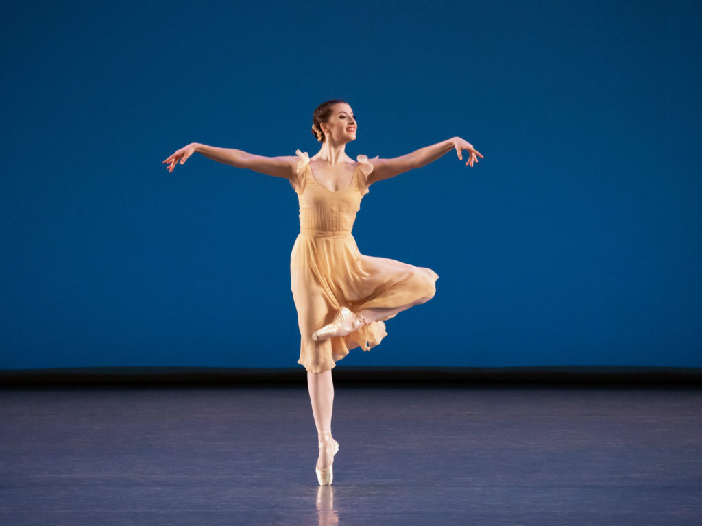 Indiana Woodward dances onstage in front of a blue backdrop and wears a cream-colored dress, pink tights and pointe shoes. She performs a relevé on her right foot and lifts her left leg into retiré, looking out over her left hand, which is stretched out in front of her, and holds her right arm out to the side.