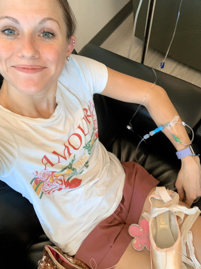 Abby Phillips Maginity takes a selfie of herself sitting in a black leather chair with an IV drip attached to her left arm. A pair of pointe shoes lays in her lap.