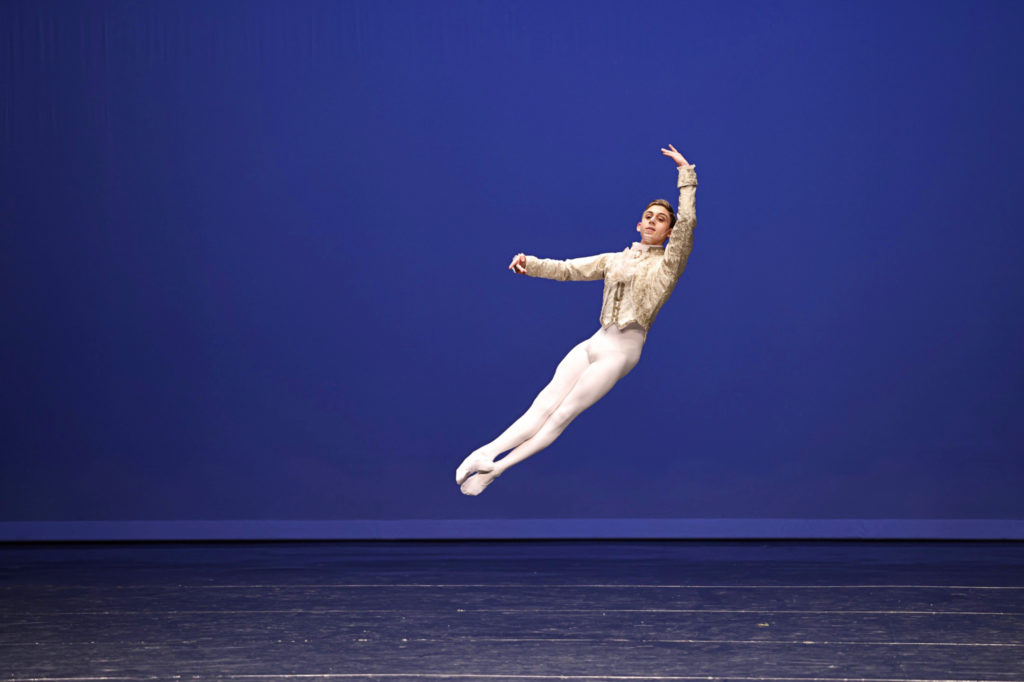 Zander Magolnick, wearing white tights and ballet slippers and a princely white and gold tunic, performs an assemblé in effacé devant onstage at a ballet competition. He lifts his left arm up over his head and his right arm is curved in front of him.