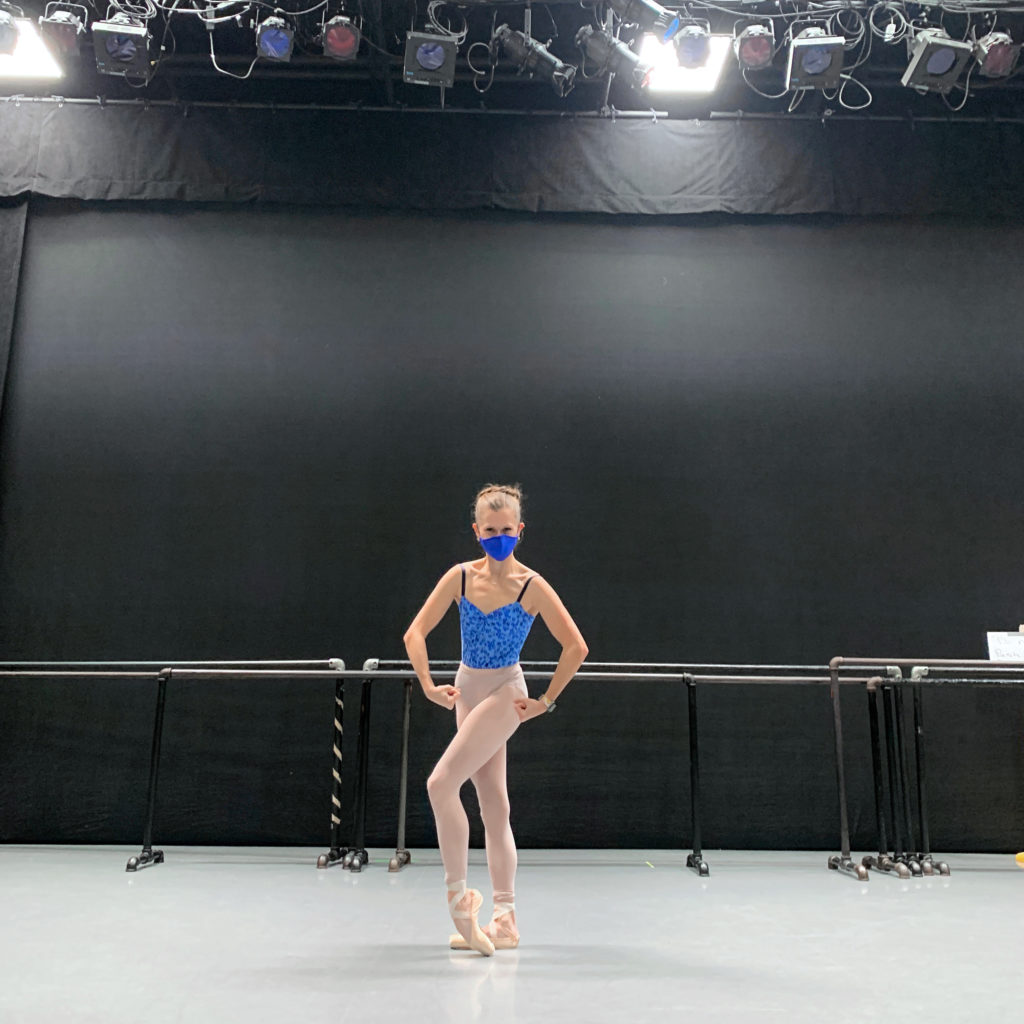 Abby Phillips Maginity wears pink tights, pointe shoes, blue face mask and a blue leotard and stands beveling her left foot on pointe. She makes a muscle-man pose with her arms while standing in a black box studio theater.