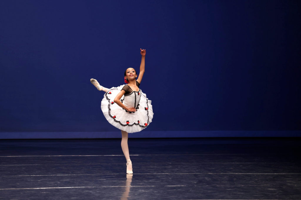 Adaline Dunlap, a young ballet student, wears a white tutu with red roses on the skirt and black trim and performs onstage. She stands on demi-pointe with her left leg in an attitude derriere croisé, and holds her waist with her right hand and her left arm up.