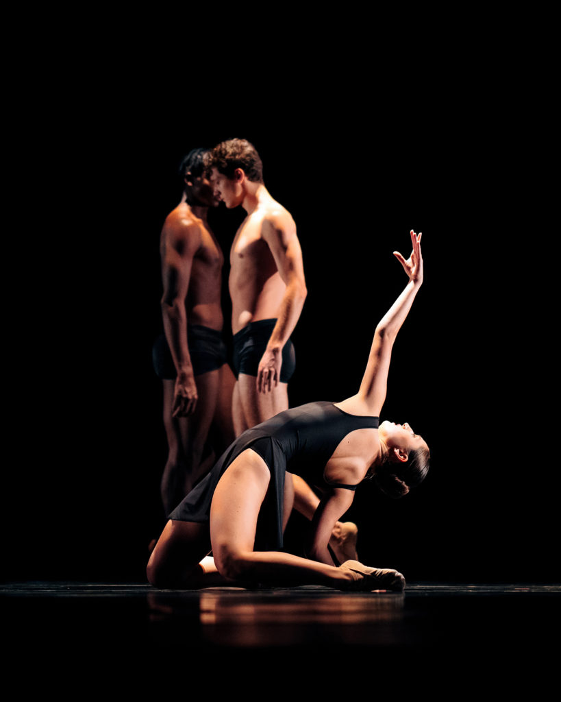 Ashlan Zay kneels on both knees onstage and bends her upper body back dramatically, touching the floor with her right hand and reaching up high with her left. She wears a black dance dress and dances in a pool of light on a dark stage. Behind her, two men in black shorts stand facing each other.