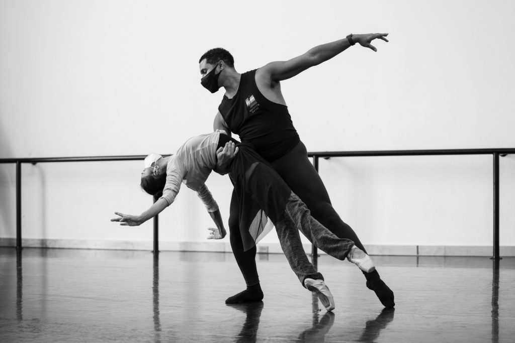 Erica De La O falls backward on pointe into a backbend, her arms pressed back like a swan, while her partner, Brandon Ragland, holds her around the waist with his right arm and tendus his left leg out to the side. They both dance in a large studio and wear layers of dance clothing.