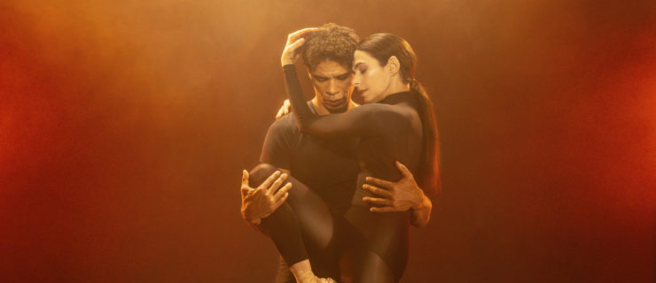 Carlos Acosta stands behind Alessandra Ferri and holds her around the waist with his left hand and her right leg up in a parallel retiré position with his right while she cradles his head in her hands. They both wear dark dance clothing and are lit with foggy, orange lighting.