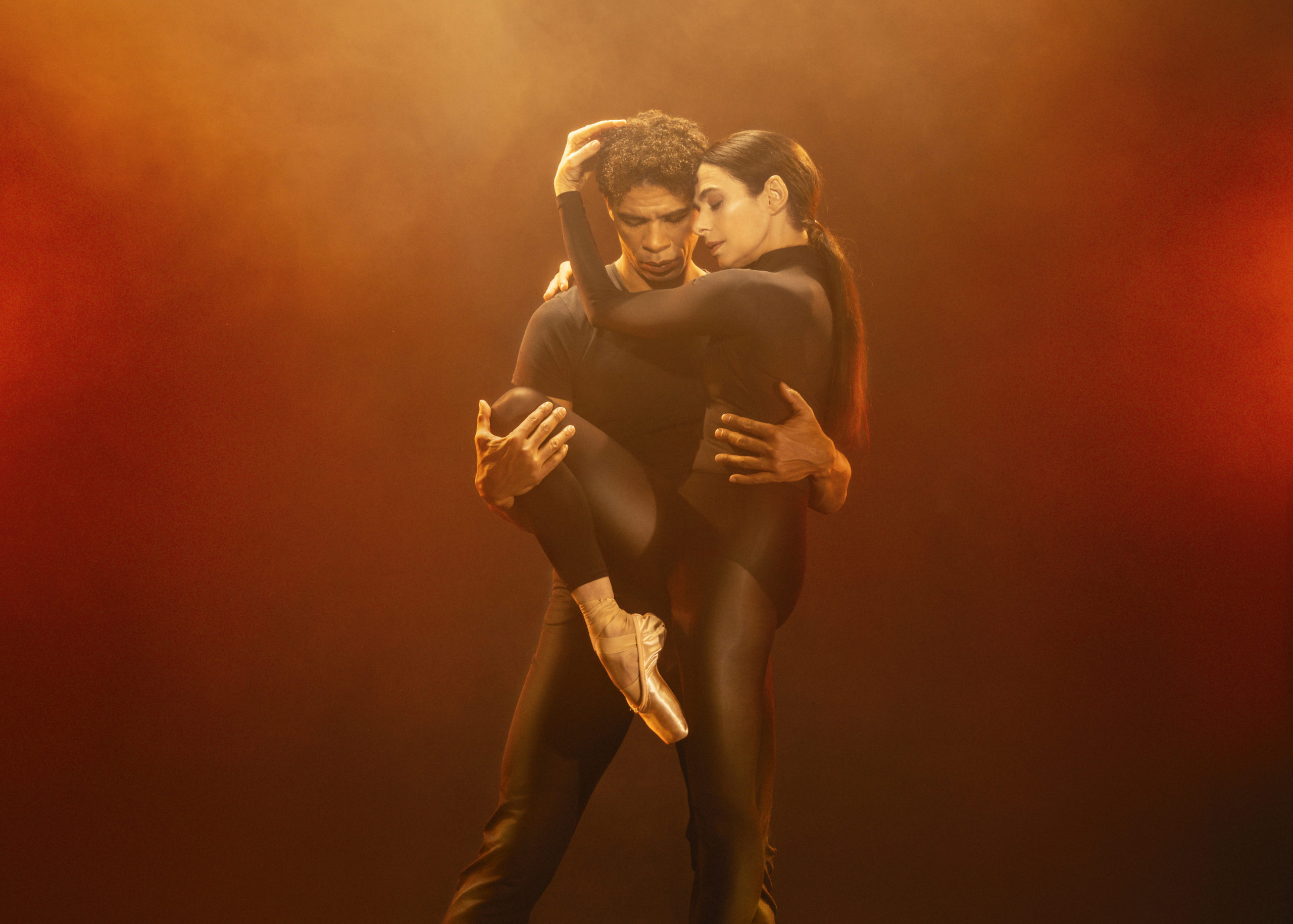 Carlos Acosta stands behind Alessandra Ferri and holds her around the waist with his left hand and her right leg up in a parallel retiré position with his right while she cradles his head in her hands. They both wear dark dance clothing and are lit with foggy, orange lighting.