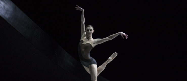 Elle Macy, wearing a dark leotard and pointe shoes, stands on her left foot on pointe on a darkened stage and kicks her right leg up behind her. She holds her right arm up high and her left arm out to the side.