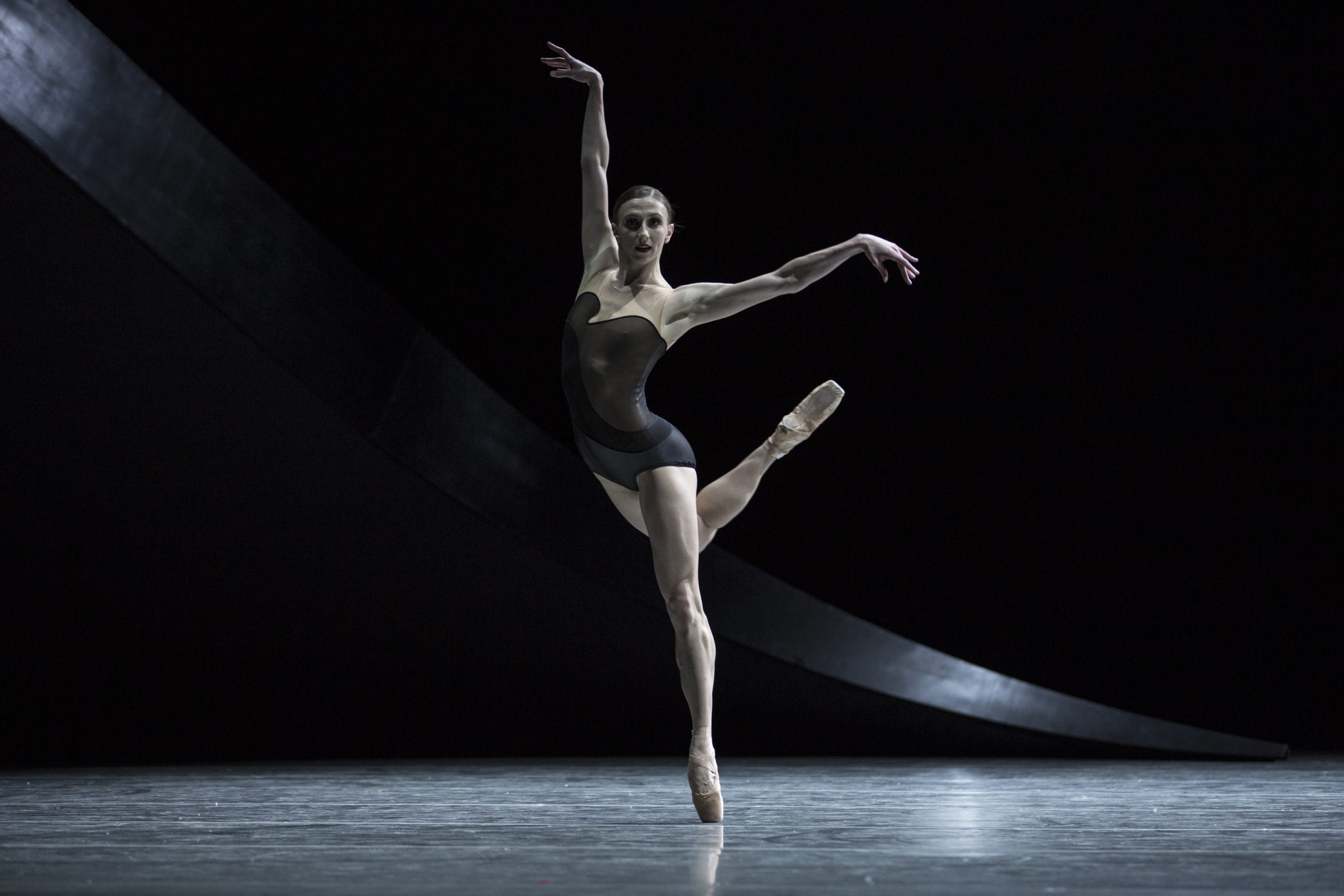 Elle Macy, wearing a dark leotard and pointe shoes, stands on her left foot on pointe on a darkened stage and kicks her right leg up behind her. She holds her right arm up high and her left arm out to the side.