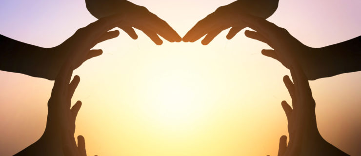 A groupe of people create a heart shape with all of their hands, shown in silhouette against a sunset.