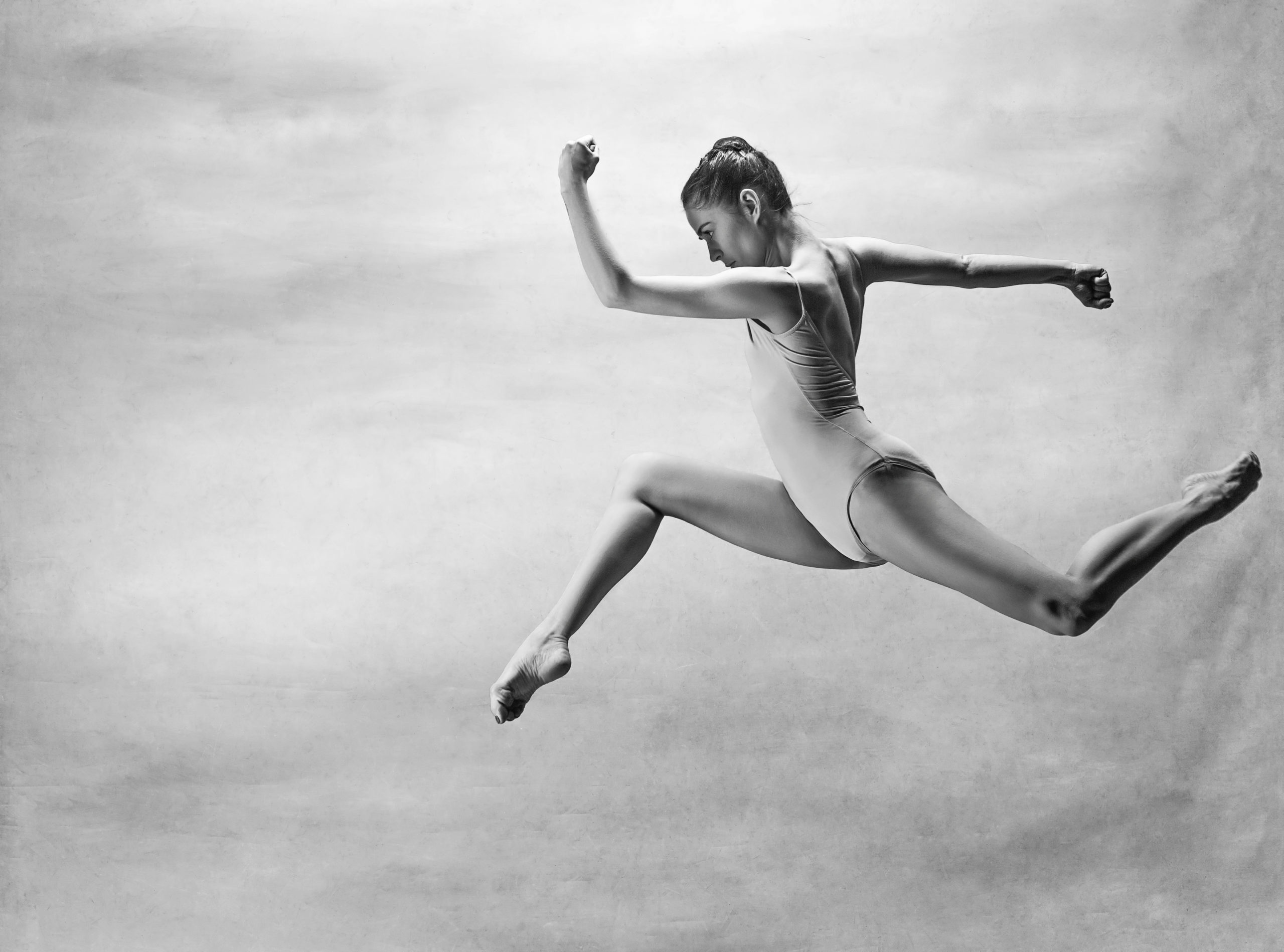 A female ballet dancer jumps up in a leap with both knees bent, with her hands in fists and her left arm crooked in front of her face. She looks determined and dances in front of a studio backdrop in this black and white image.