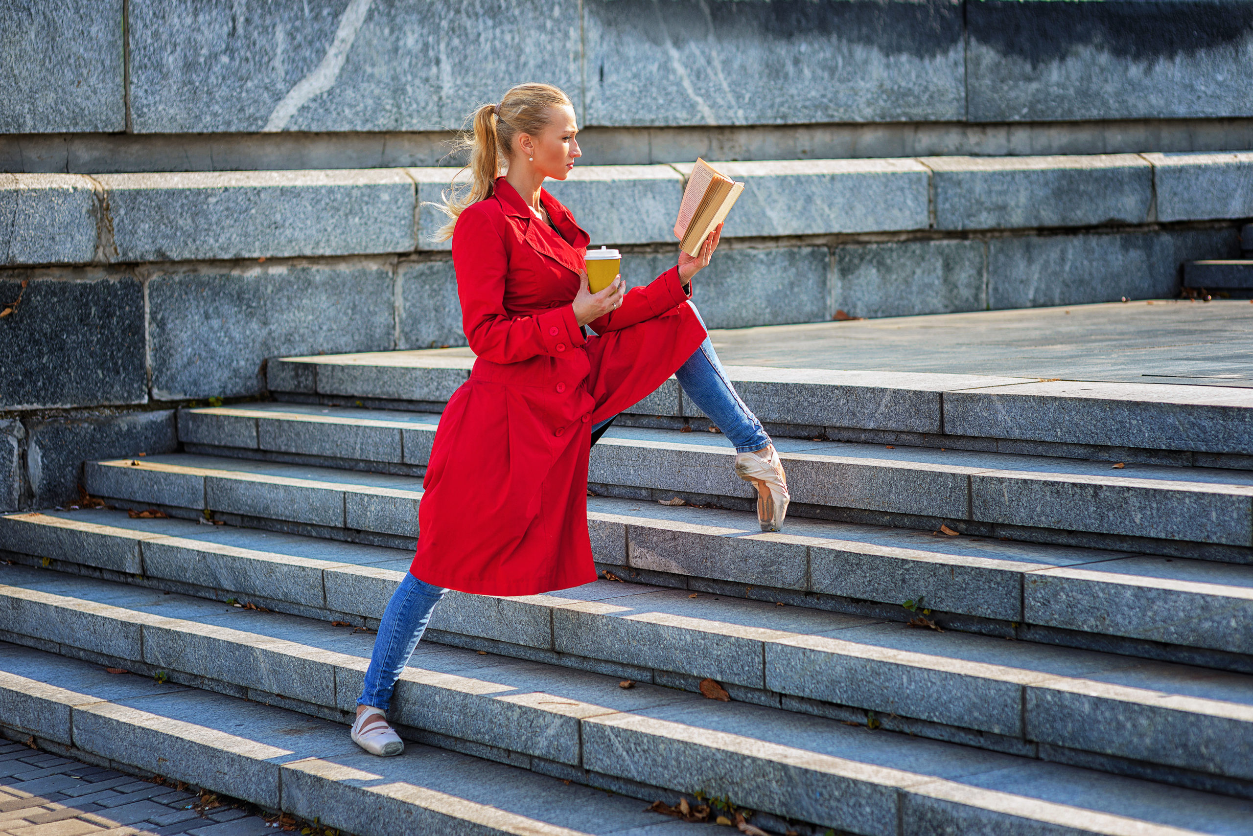 A ballerina in a red trench coat, blue jeans and pointe shoes poses on an outdoor staircase, propping her left leg on pointe three stairs above her right leg. She holds a cup of coffee in her right hand and a book, which she is reading, in her left.