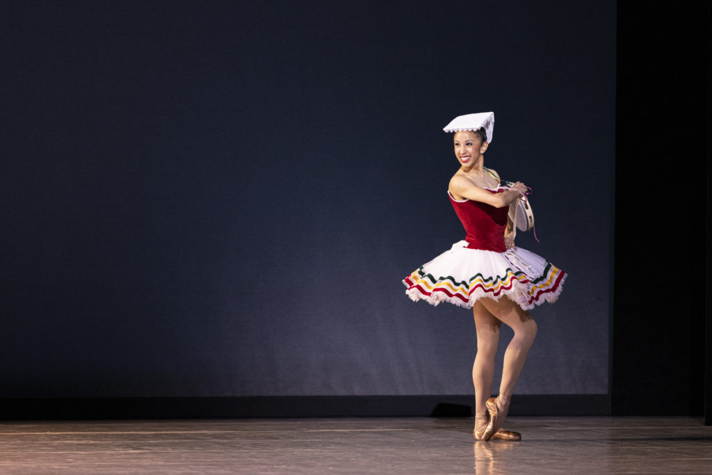 Jenna Rae Herrera stands onstage, her body facing the wings, and looks out to the audience with a huge smile. She pops her right foot up onto point and holds her left hand to her waist while pulling a tambourine close to her chest with her right hand. She wears a tutu with a red bodice and white skirt with red, green and yellow stripes trimming the edge and a white, square-shaped headpiece.