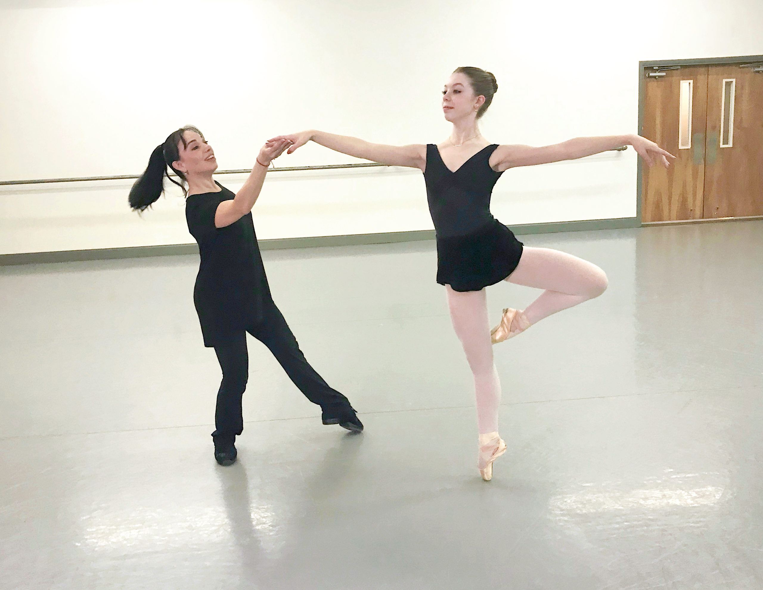 Katia Garza, wearing black pants and a long T-shirt, holds McKinley White's right hand as she practice a piquè turn manège. Mckinley wears a black leotard and ballet skirt, pink tights and pointe shoes, and they practice in a large dance studio.