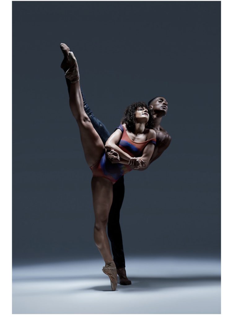 Brandon Gray stands behind Larissa Gerske, reaching around to hold her hands as they both execute a grand battement in second. She wears an orange and blue striped leotard and brown pointe shoes, while he wears dark tights and ballet slippers.