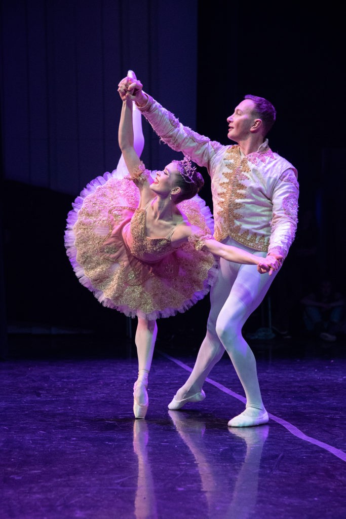 A ballerina in a pink tutu with gold embellishments and a sparkly crown performs a penché on pointe while her partner, in a white prince costume, stands behind her and holds her hands. They perform onstage in The Nutcracker and smile toward the audience.