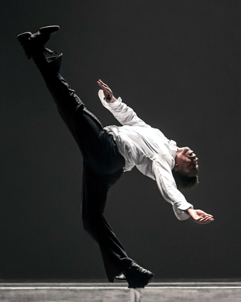 Maurice Mouzon, wearing a white button down shirt, black pants and black dance sneakers, performs a dramatic layout with his left leg extended high.
