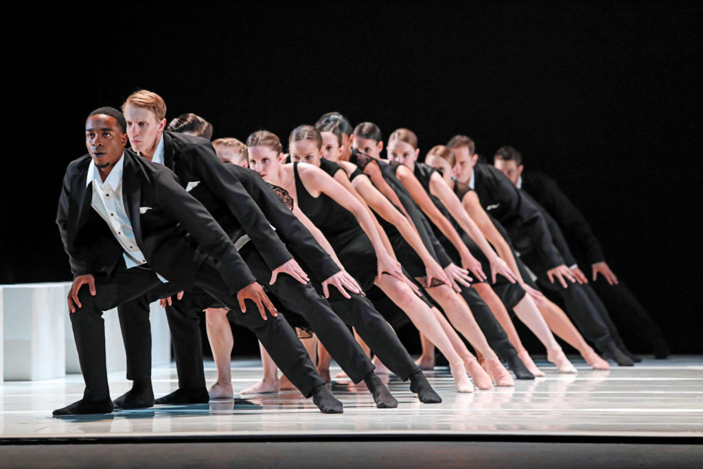 A line of both male and female dancers create a singel file line onstage and pose in a wide stance, hands on thier knees, and look out towards the audience. The men wear black cuits with white shirts and black socks, and the women wear knee-length black dresses and pick ballet shoes. They wear intense facial expressions, without smiles.