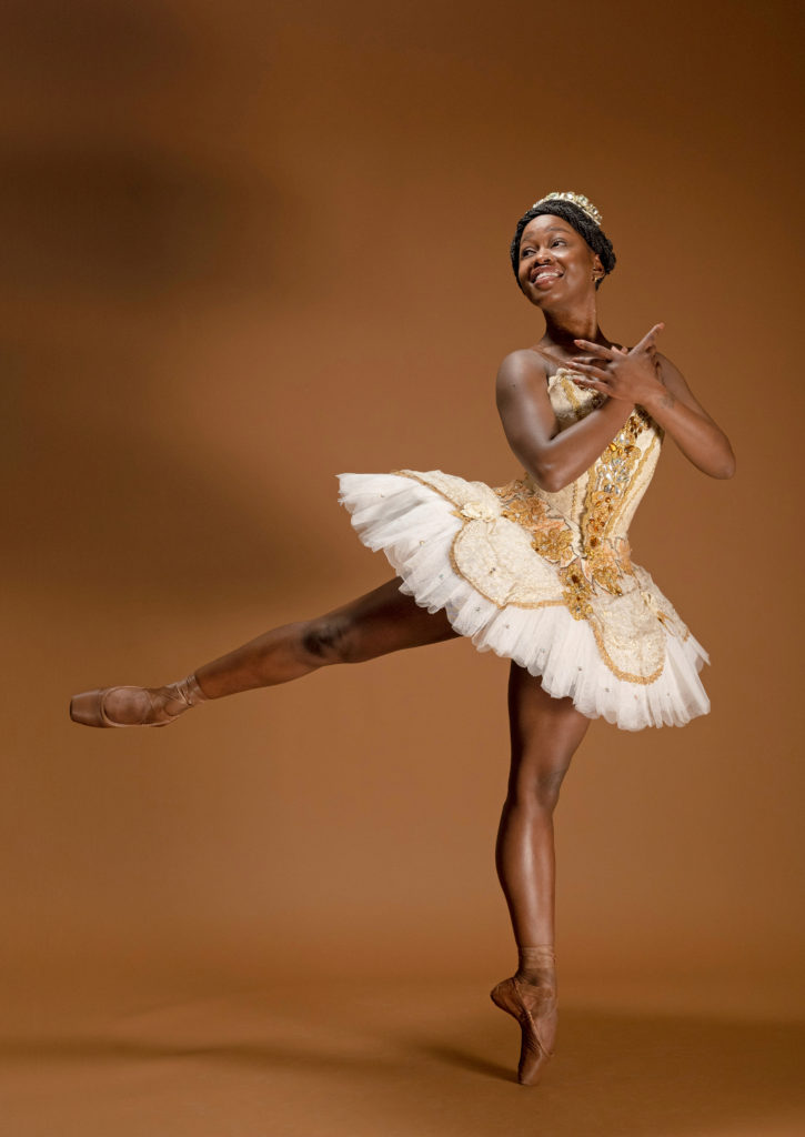 Michaela DePrince poses in a low arabesque effacé on pointe, her right leg lifted, and her arms crossed at her chest. She wears brown pointe shoes, a white and gold classical tutu and crown, with her braided hair parted down the middle and in a low bun.