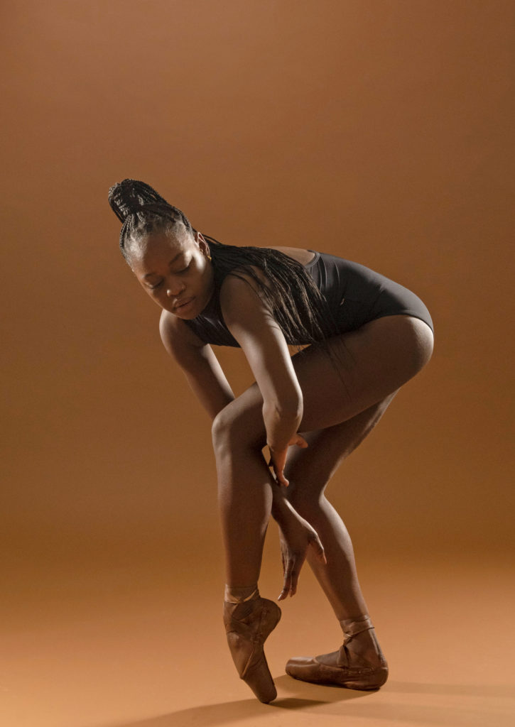 Michaela DePrince stands in profile and bends low with her left leg popped up onto pointe. She reaches her arms down alongside her left calf and looks towards the floor. She stands in front of a light brown backdrop and wears a black leotard, brown pointe shoes and her braided hair in a high ponytail.