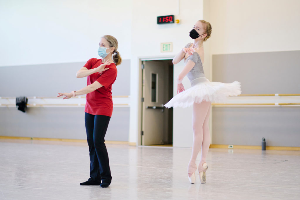 Tina LeBlanc wears a red shirt and black pants and moves her arms across her body in a large dance studio. Behind her, Sasha De Sola wears a white practice tutu and stands in sus-sous on pointe, brining her right hand in towards her left shoulder and her left arm across her waist.