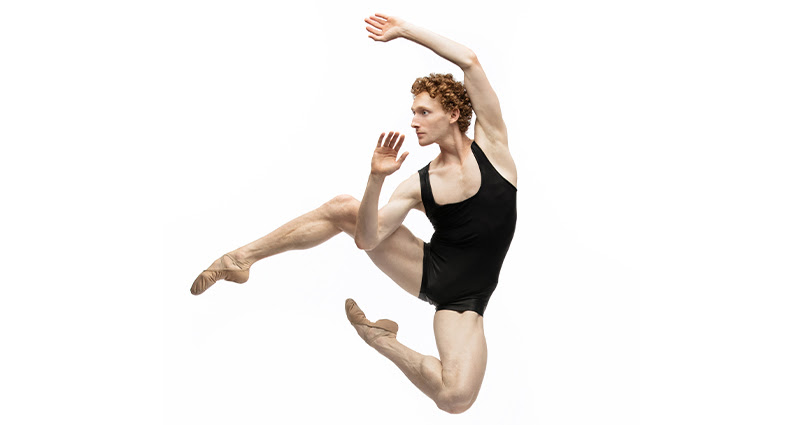 Against a white backdrop, Skylar Campbell, wearing a black tank top and bike shorts, jumps up with his right leg out to the side, both legs bent. He lifts his left arm above his head and bends his right arm near his body so that his hand is close to his face.