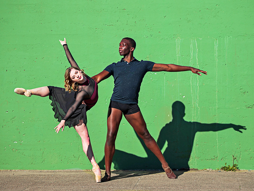 Cassidy Isaacson poses in a croise attitude on pointe while Brandon Alexander holds her waist and leans her out so that she makes a dramatic angle with her body. They pose outdoors on the pavement in front of a bright green wall; Cassidy wears a black mesh dance dress with a pink front and Brandon wears black booty shorts, a blue polo shirt and brown ballet slippers.