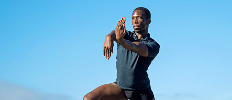 Brandon Alexander lunges dramatically outdoors in an arid landscape. He pops his right foot up onto demi-pointe and flexes his hands in front of him, wearing a blue polo shirt and black booty shorts.