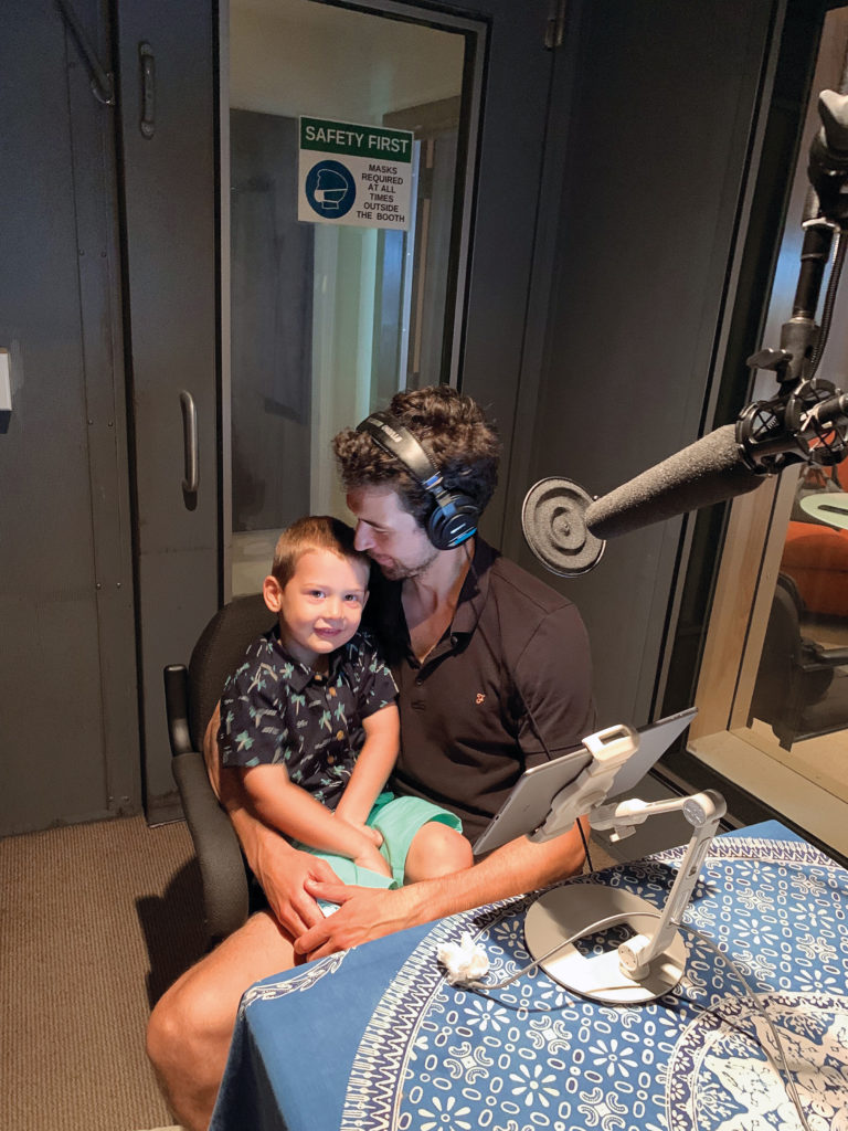 During a podcast recording, Thomas Forster sits wearing headphones in a small room holding his young son on his lap. They both wear short sleeved shirts and shorts.