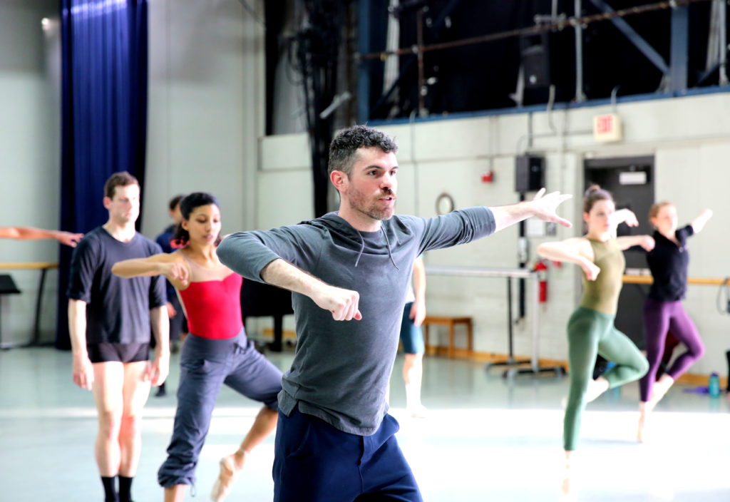 Tom Mattingly, wearing a gray sweatshirt and blue pants, demonstrates a move with his arms to a large studio full of dancers.