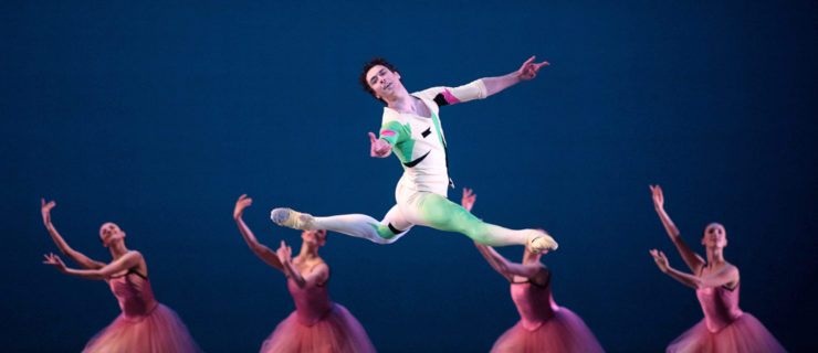Thomas Forster performs a Bournonville-style grand jeté onstage, looking out towards the audience with a smile. He wears a white shirt and tights with green, black and pink shapes on them and ballet slippers. Behind him a line of women in pink tutus kneel on their left knee with their right leg extended forward.