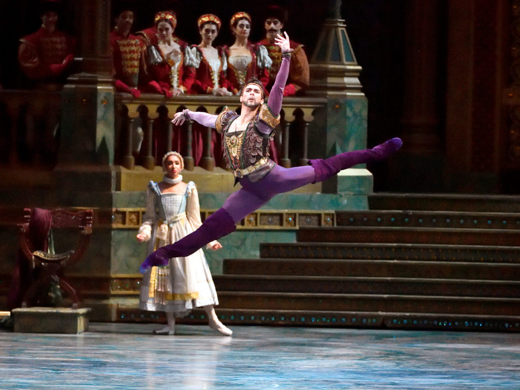 Wearing a purple and gold jackaet, purple tights and sued purple ballet boots, Thomas Foster performs a large sissone with split legs. Behind him is a grand staircase and a corps of women in medieval costumes watching him.