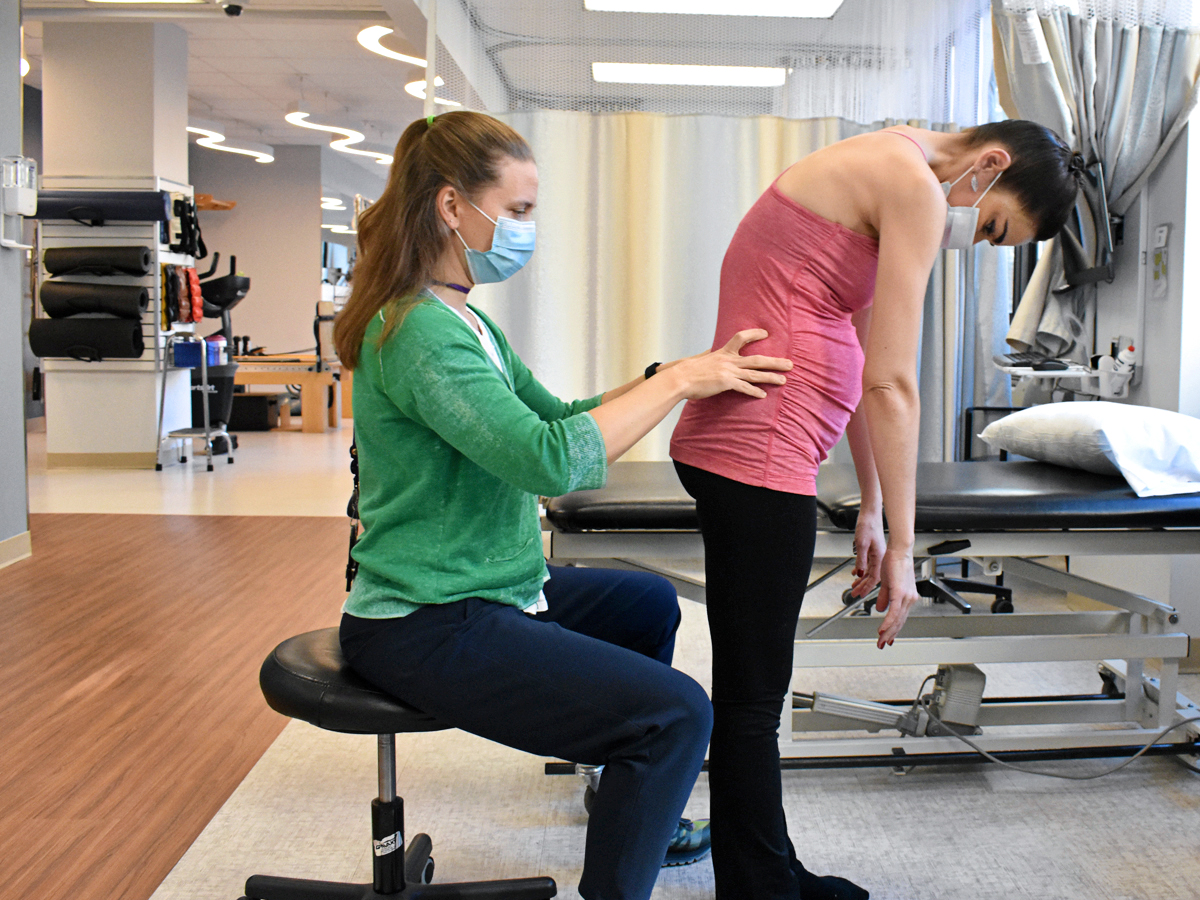 Heather Heineman, sitting on a stool and wearing a green sweater and blue pants, places her hands on the lower back of a pregant Kate Loh, who bends over from the mid-back. Loh wears a pink athletic tank top and black yoga pants, and the two work in a physical therapy clinic.
