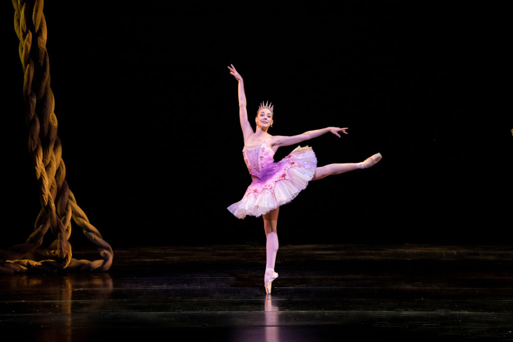 Jaime DeRocker performs a first arabesque onstage and looks out towards the audience with a large smile. She wears a pink tutu and tiara, pink tights and pointe shoes.