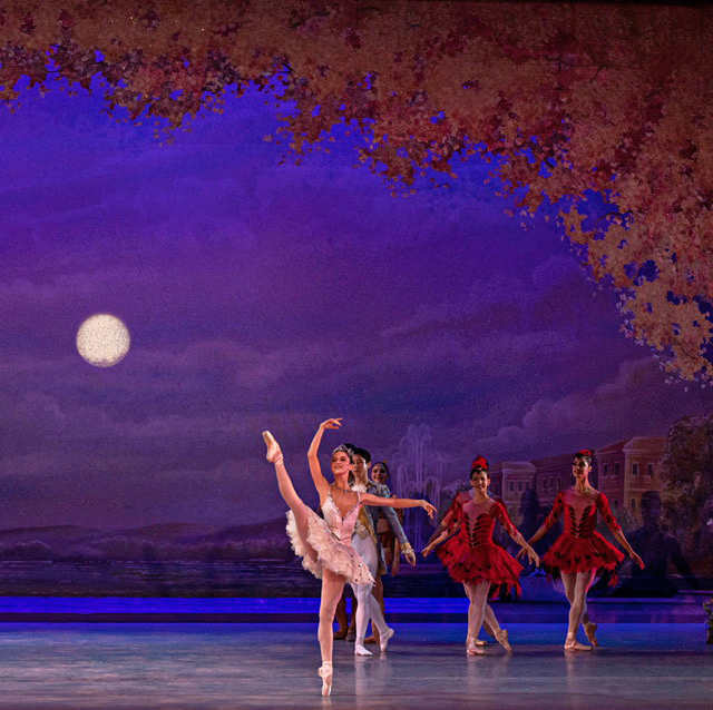 Adelaide Clauss, in a pink tutu, pink tights and pointe shoes, performs onstage during a performance of the Nutcracker. She does a développé à la seconde with her right leg and holds her right ar above her head. A line of dancers stand in B+ behind her.