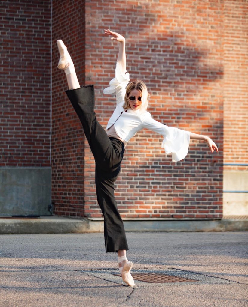 Sage Humphries, wearing sunglasses, a fashionable white blouse, black pants and pointe shoes, performs a grand battement in croise devant on pointe. She extends her right arm up and her left arm out to the side, and dances outside on the sidewalk in front of a brick wall.