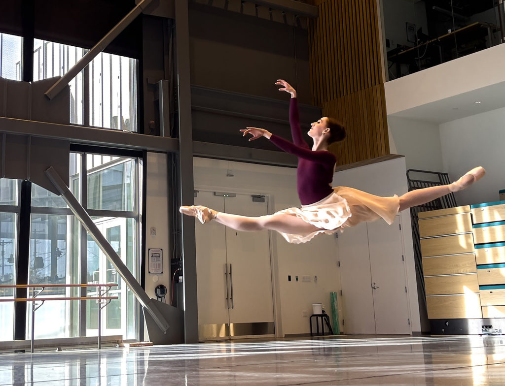 Beth Ann Maslinoff leaps trhough a bright dance studio. She wears a leotard and a mid-length ballet skirt. 