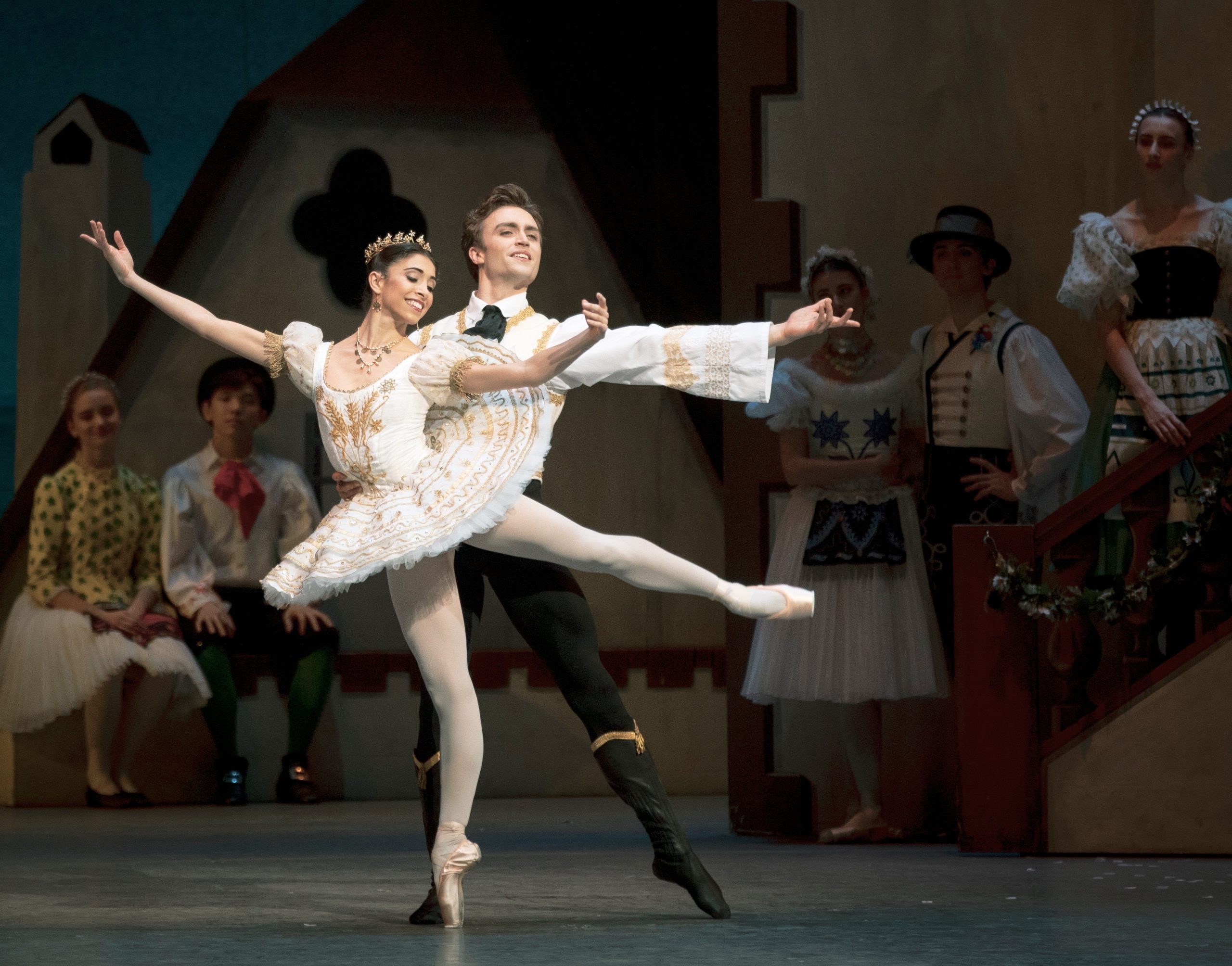 Yasmine Naghdi wearing a white tutu, tiara, pink tights and pointe shoes, poses in a low arabesque and opens her arms to second position. Behind her, Matthew Ballet, in a white shirt, black tights and black ballet boots, holds her waist and points his left foot out in tendu, and holds his left arm out in second. They dance onstage during a village scene, and dancers dressed as townspeople stand behind them watching them dance.