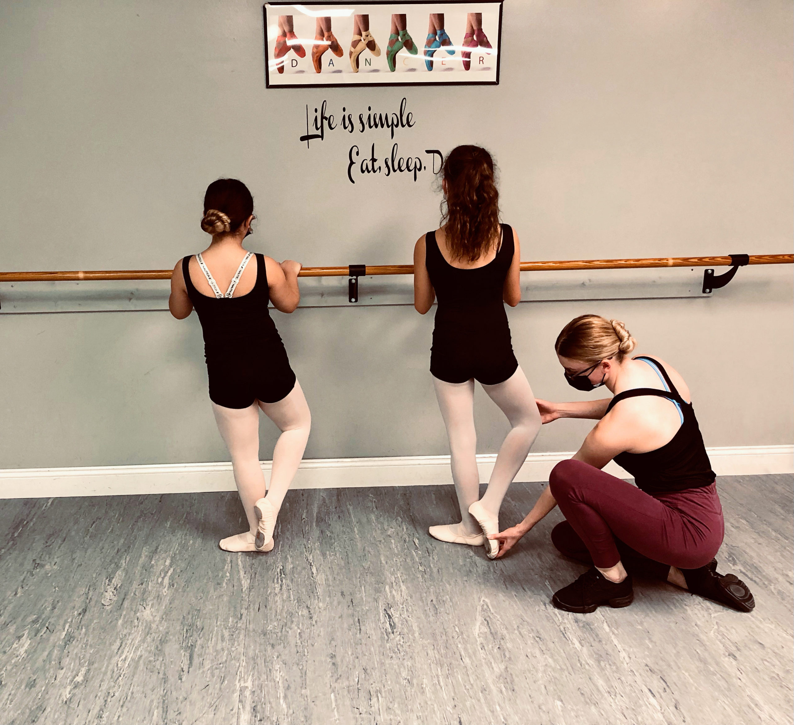 In a small dance studio, two little girls are shown at the barre practicing coupé derriere with their right foot. Their ballet teacher, wearing a black face mask, black leotard and burgundy leggings, crouches down to corrects the foot of the girl on the right.
