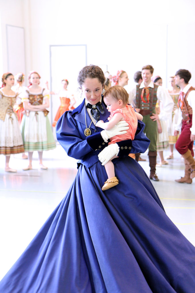 Sarah Ricard Orza stands in a dance studio wearing a blue 19-century style dress. She twrils while holding her young infant daughter. Behind her, other dancers in costumes stand and talk casually.