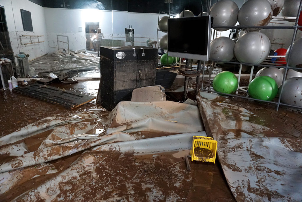 A dance studio is shown in shambles, with the studio floor covered in water and mud, with scattered debris and a door off its hinges on the ground.
