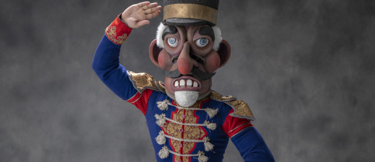 A male dancer is shown from the waist up wearing a Nutcracker costume and large Nutcracker head. He holds his left hand at his waist and salutes with his right hand.